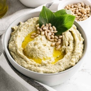 A bowl of pesto hummus with olive oil and pine nuts sprinkled on top.