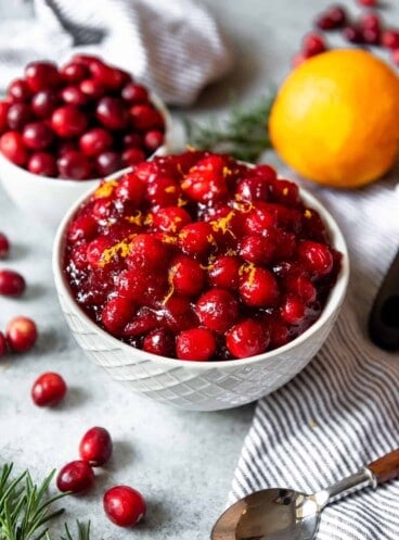A white bowl filled with cranberry sauce with orange zest in it, next to a bowl of cranberries, a spoon and an orange on a festive table.