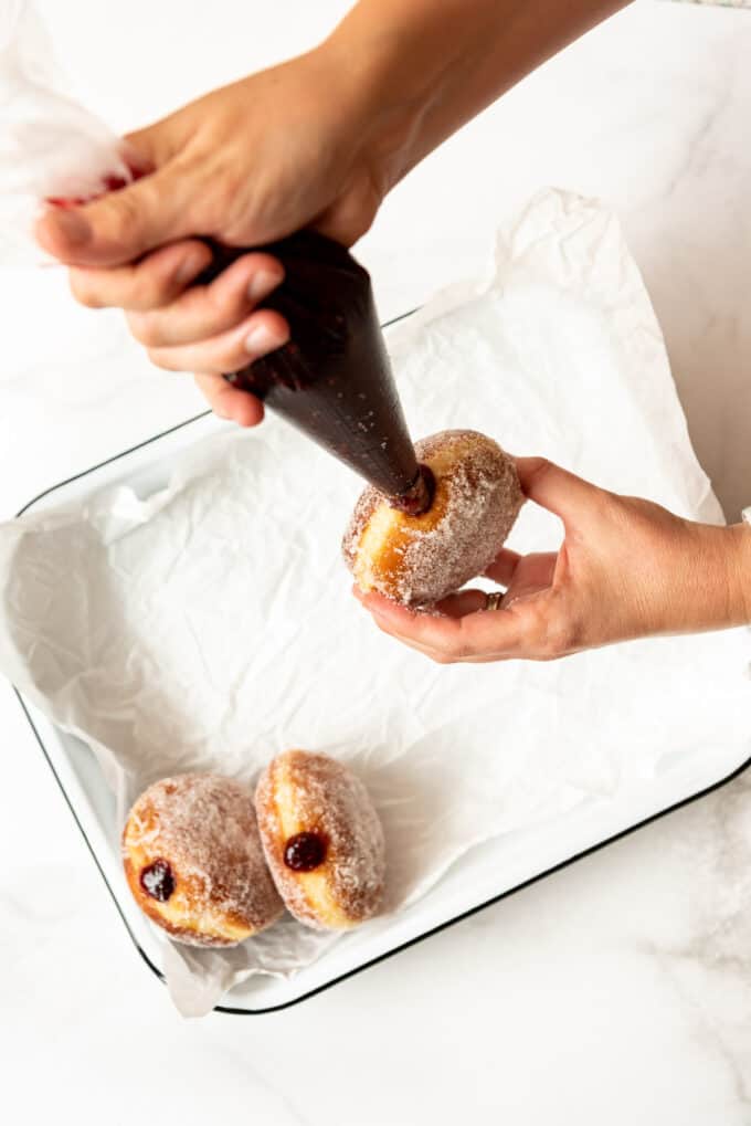 Hands filling a homemade packzi donut with raspberry jelly.