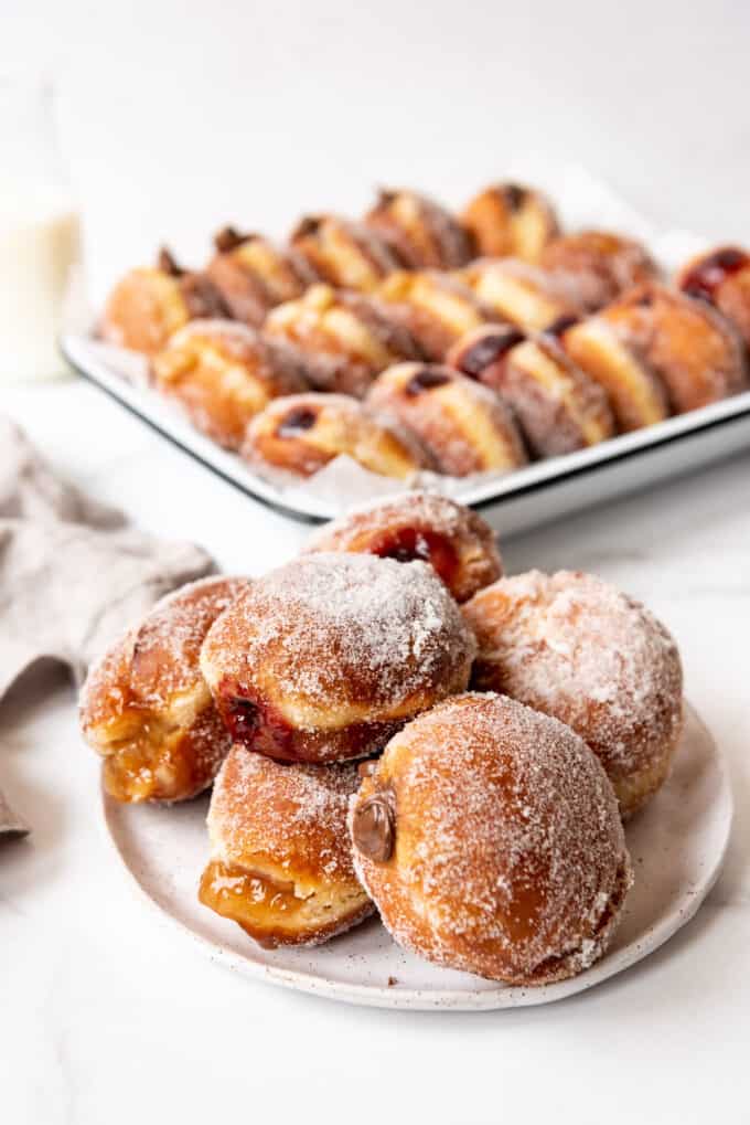 A pile of packzi donuts on a plate in front of more donuts stacked in rows in a pan.
