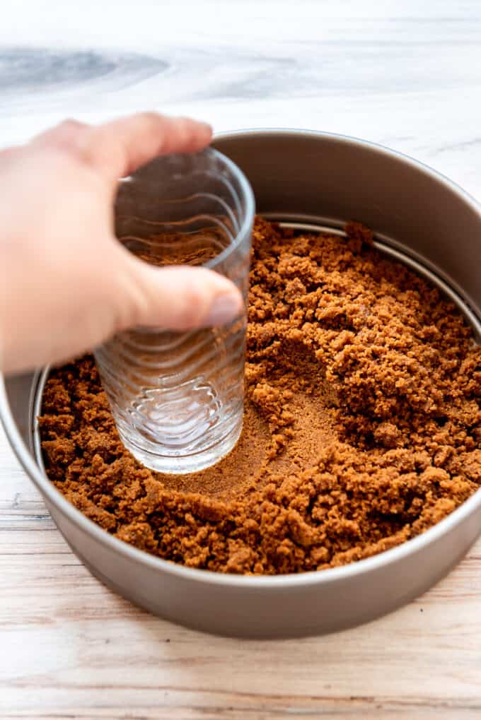 A hand holding a glass to press cookie crumbs into a springform pan for a cheesecake crust.