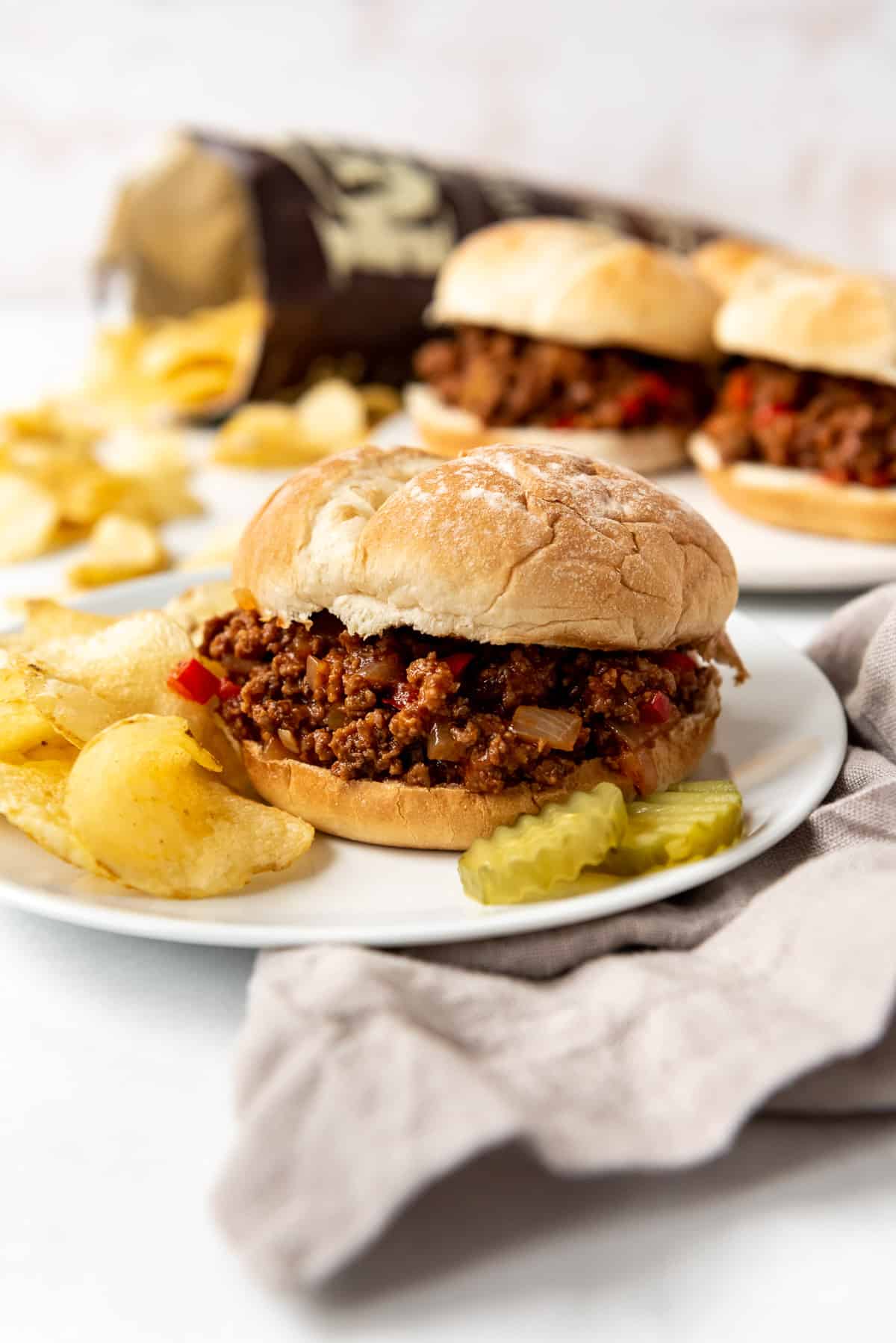 A sloppy joe sandwich on a plate next to a linen napkin and in front of a bag of chips and more sloppy joe sandwiches.