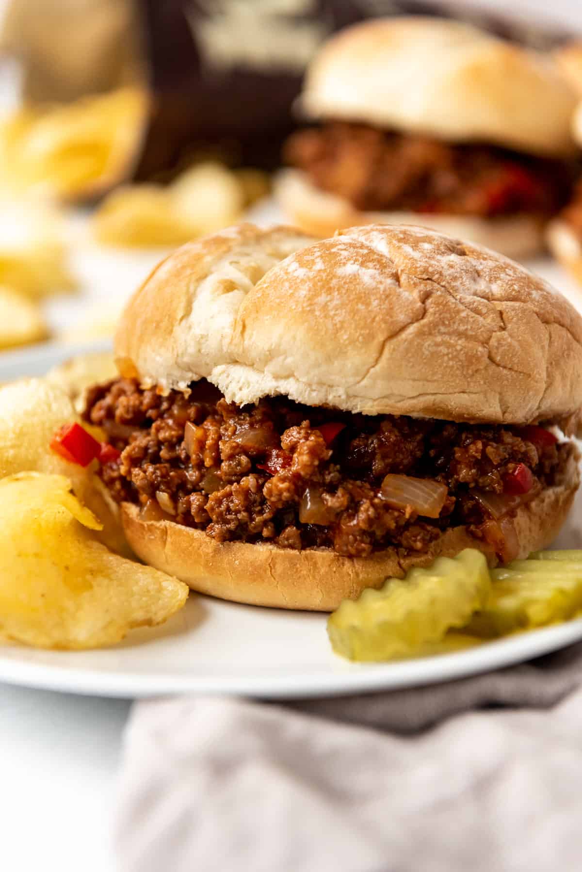 A homemade sloppy joe sandwich on a plate with pickles and potato chips.