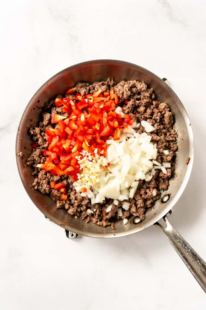 Adding onions, garlic, and red peppers to browned ground beef in a pan.