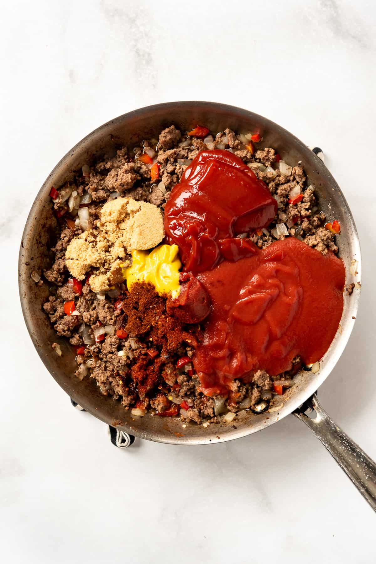 Adding ketchup, tomato sauce, brown sugar, mustard, and spices to a pan full of browned ground beef.