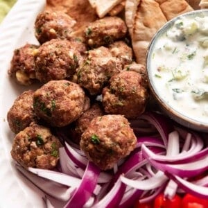 Baked Greek meatballs on a plate next to tzatziki sauce and red onions.