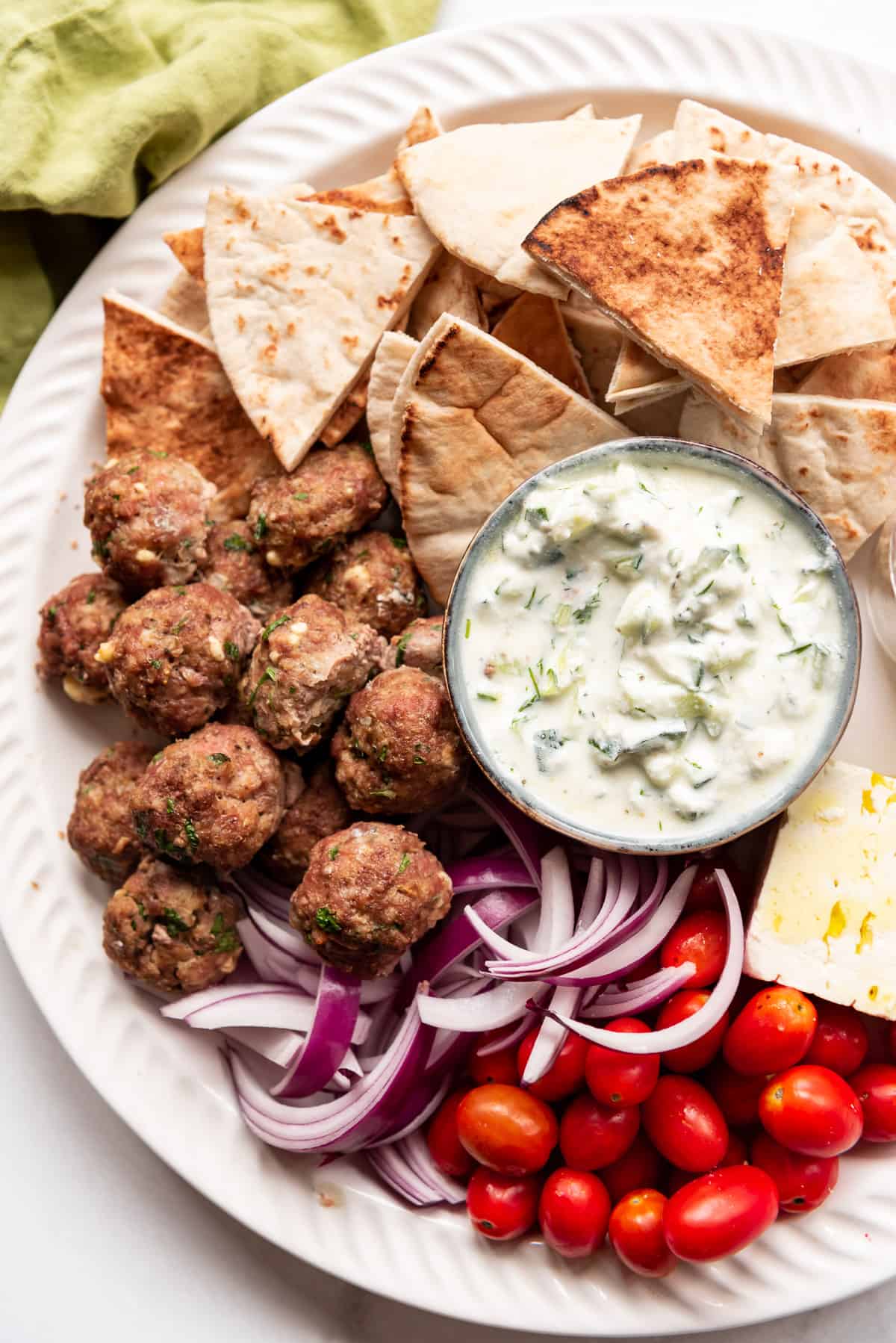 A Greek meze platter with meatballs, tzatziki sauce, pita bread triangles, red onions, grape tomatoes, and feta cheese.