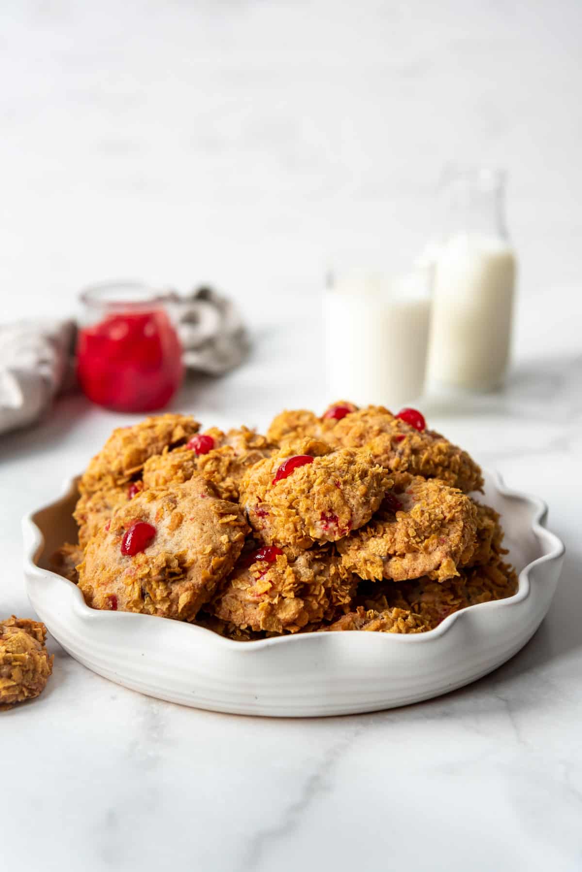 Cherry, date, and pecan cookies with crushed cornflakes on the outside in front of glasses of milk.