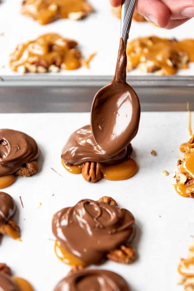 A spoon being used to spread melted chocolate onto caramel covered pecans.
