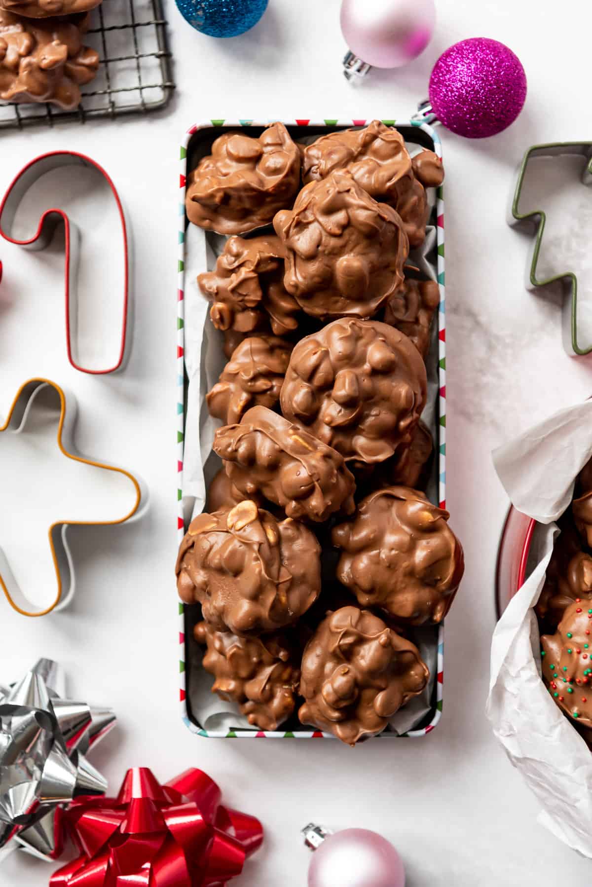 An overhead image of a rectangular tin filled with Christmas crockpot candy surrounded by ornaments, bows, and cookie cutters.
