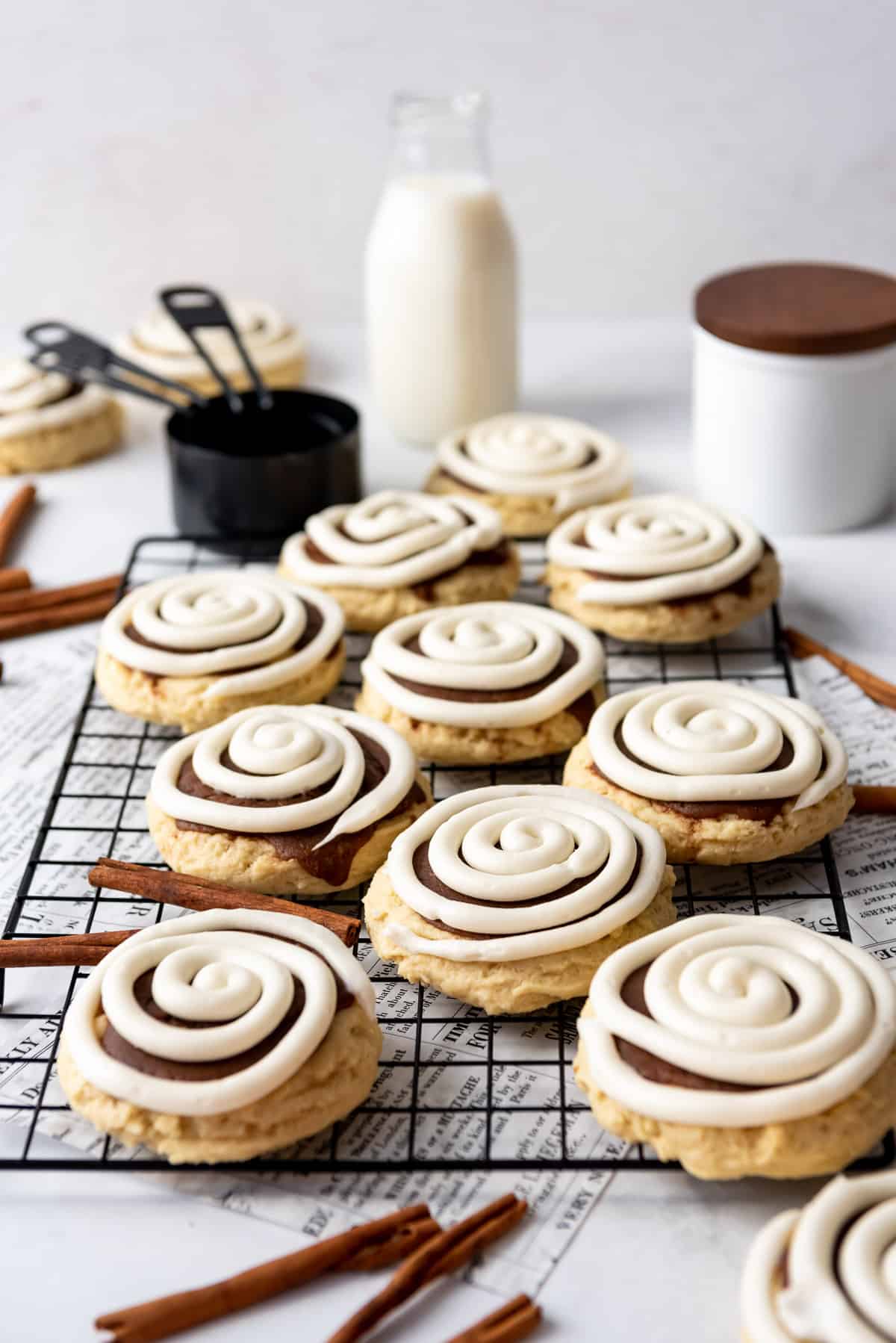 Large cinnamon roll cookies with swirls of frosting on top on a black cooling rack with a glass of milk and measuring cups behind them.