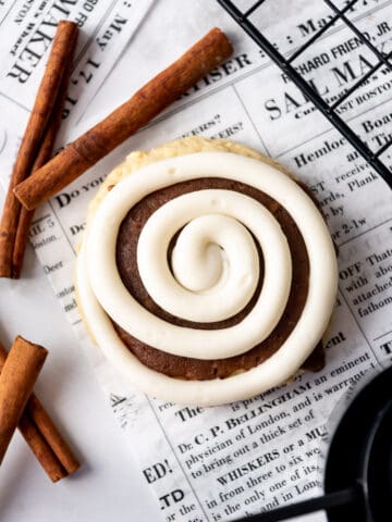 A cinnamon roll cookie with a swirl of cream cheese frosting piped on top sitting on newsprint.