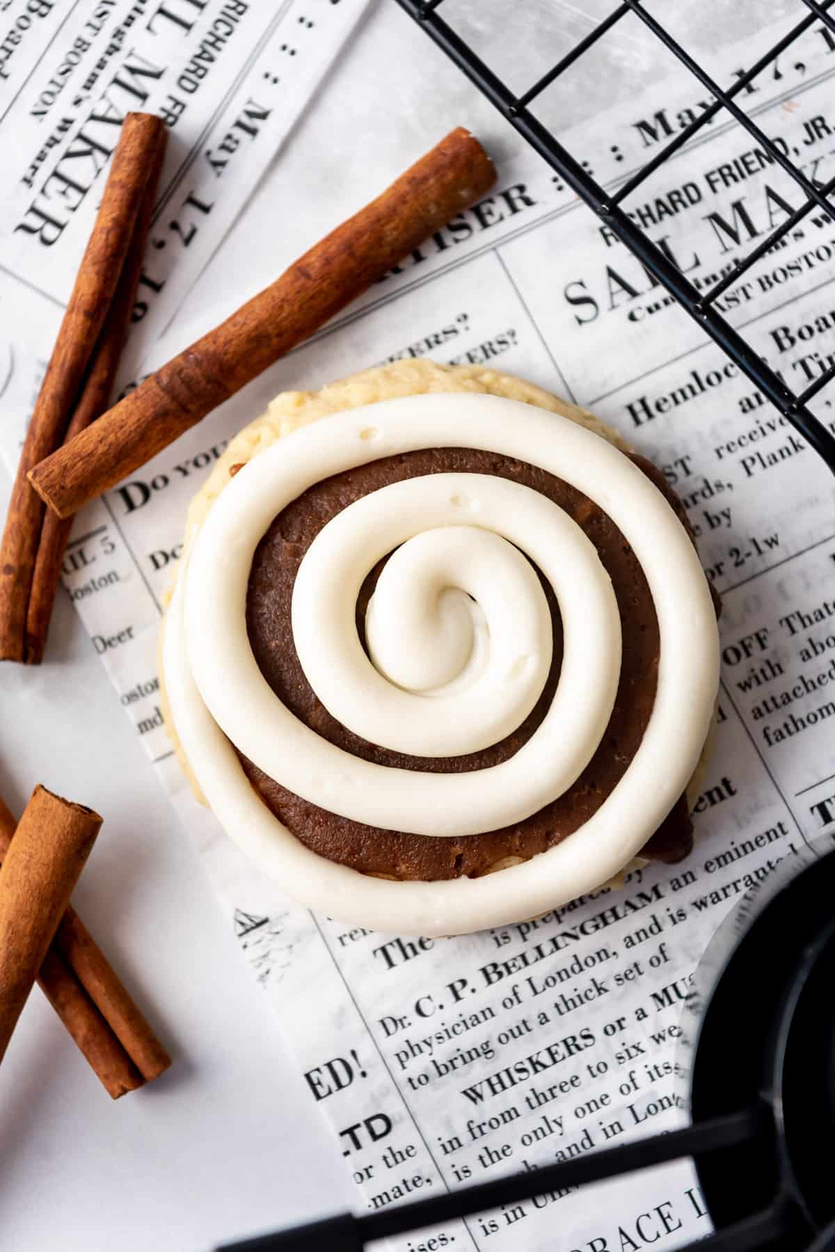 A cinnamon roll cookie with a swirl of cream cheese frosting piped on top sitting on newsprint.