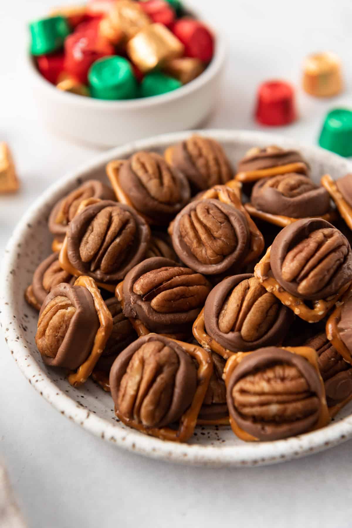 Rolo pretzels candy with pecans arranged on a plate with a bowl of wrapped Rolos behind it.