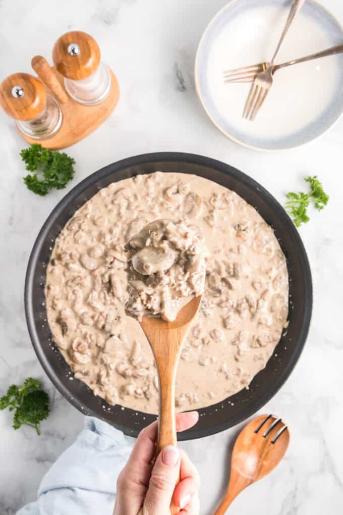 A hand lifting up a spoonful of ground beef stroganoff over a panful of the dish.