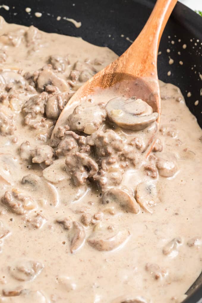 A wooden spoon stirring beef stroganoff in a cast iron skillet.