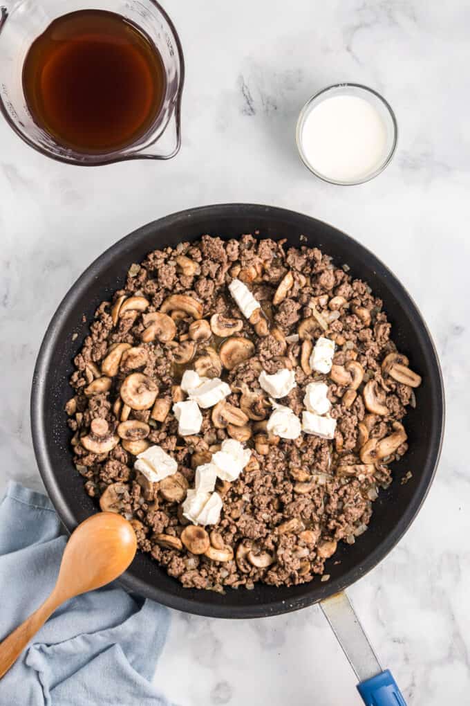Pieces of cream cheese melting over hot ground beef and sauteed mushrooms in a large cast iron pan.
