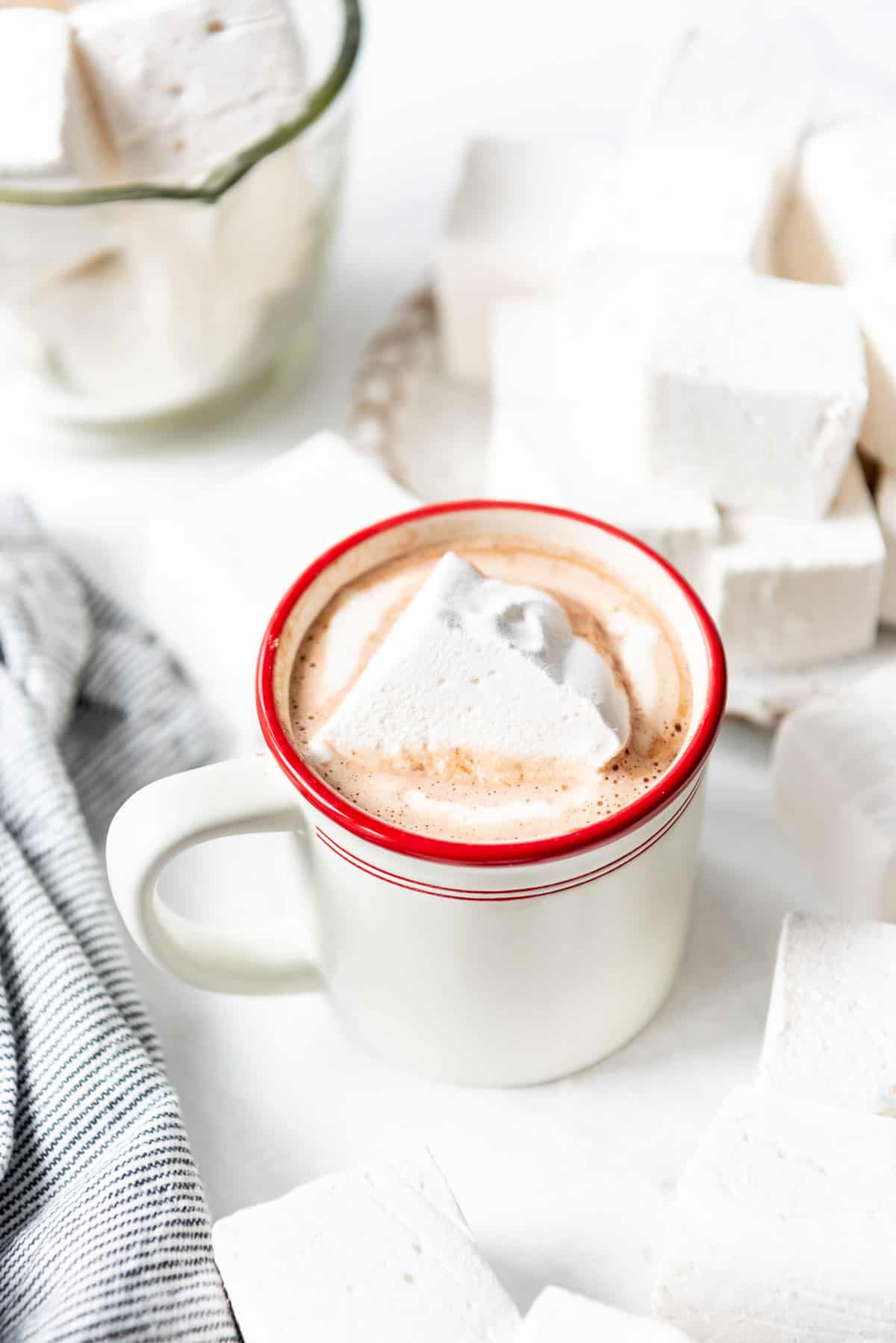 A large homemade marshmallow in a mug of hot chocolate with a red rim surrounded by more large marshmallow cubes.