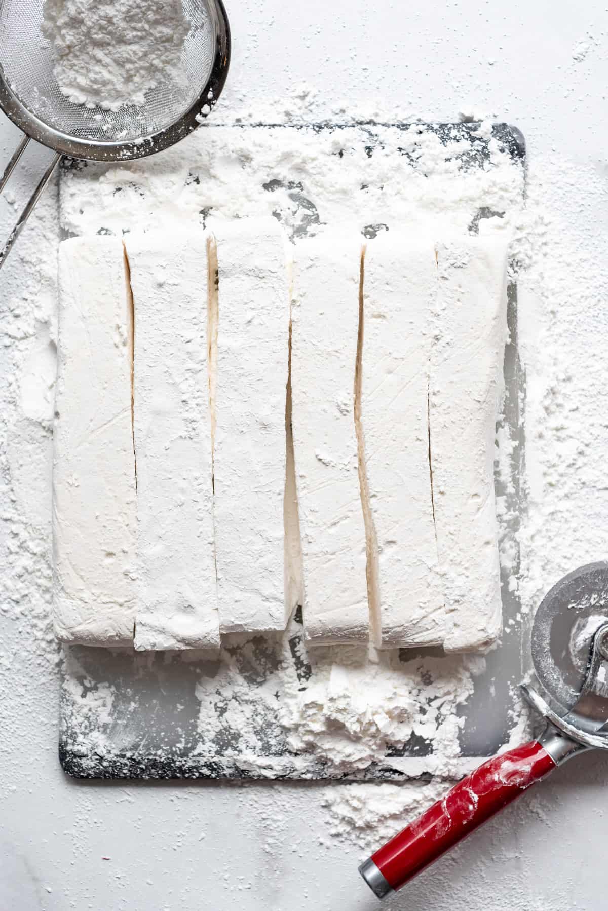 Six freshly cut rows of homemade marshmallow on a cutting board next to a pizza cutter and fine mesh sieve with powdered sugar and cornstarch.
