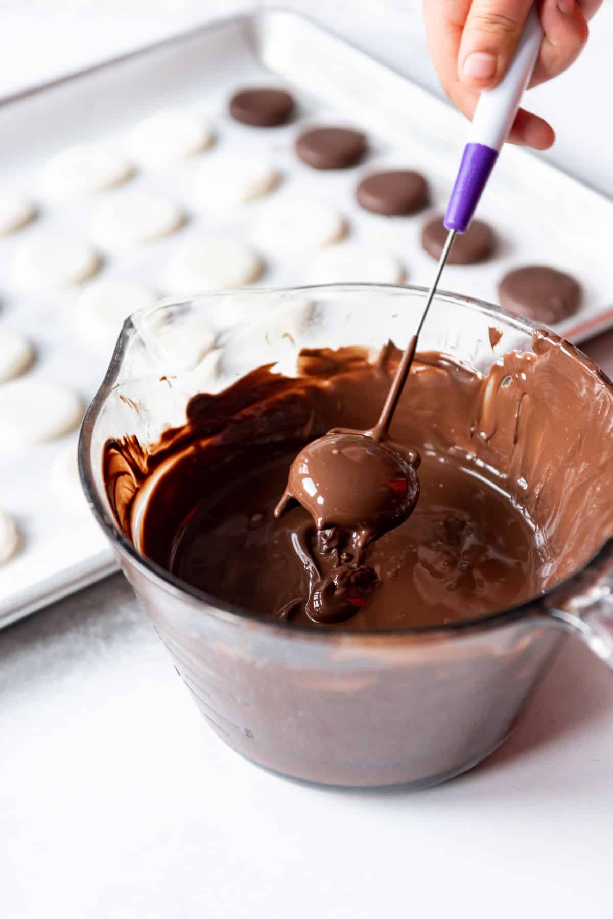A chocolate dipping tool holding a recently dipped peppermint patty center over a bowl of melted chocolate.