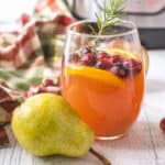 A glass of apple & pear cider made in the instant pot garnished with cranberries, orange slices, and rosemary next to a pear.