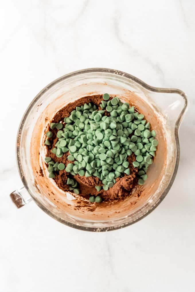 Adding green mint chips to chocolate cookie dough in a glass bowl.