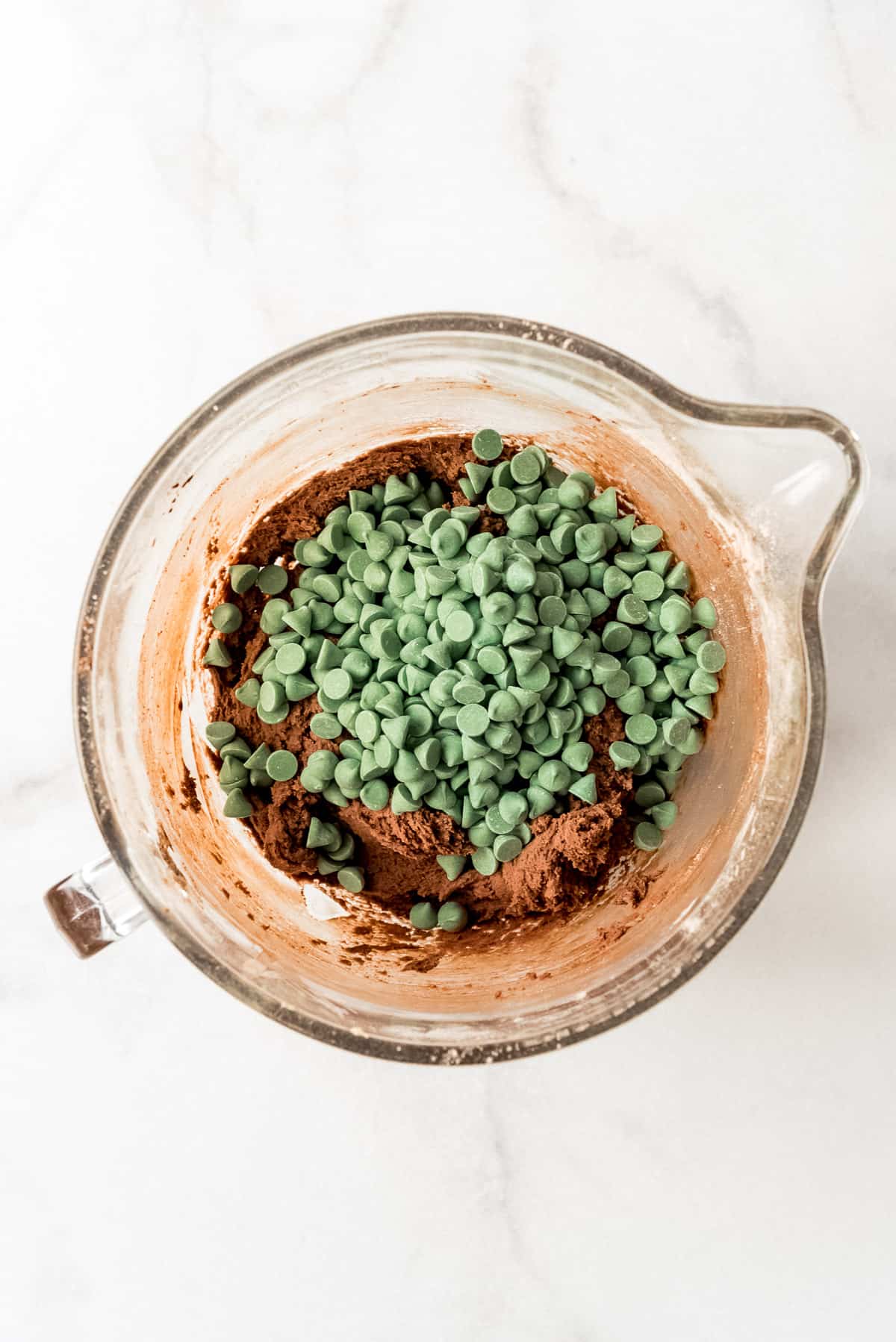 Adding green mint chips to chocolate cookie dough in a glass bowl.