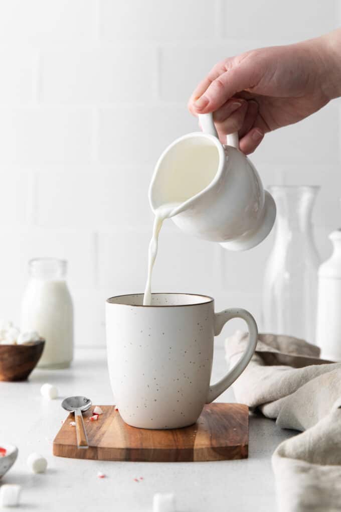 A hand pouring hot milk from a white pitcher over a hot cocoa bomb in a white mug.
