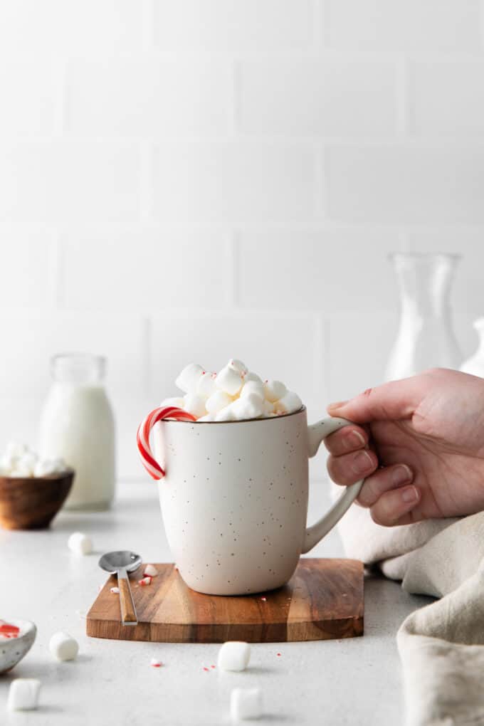 A hand lifting up a cup of hot chocolate topped with marshmallows and a candy cane.