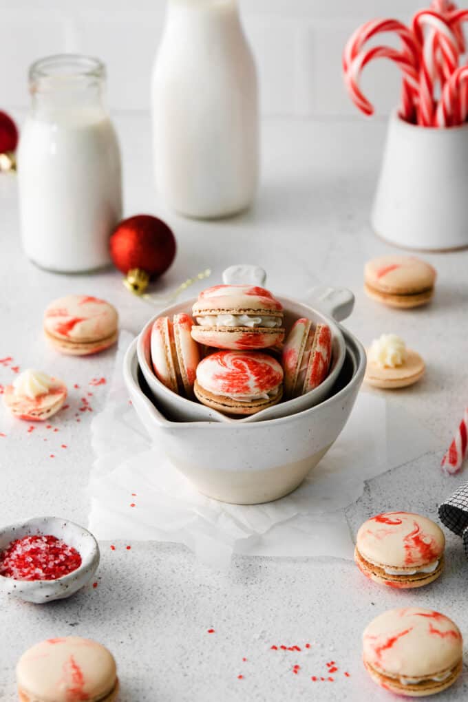 Peppermint swirl macarons displayed in nestled bowls with glasses of milk and candy canes in the background.