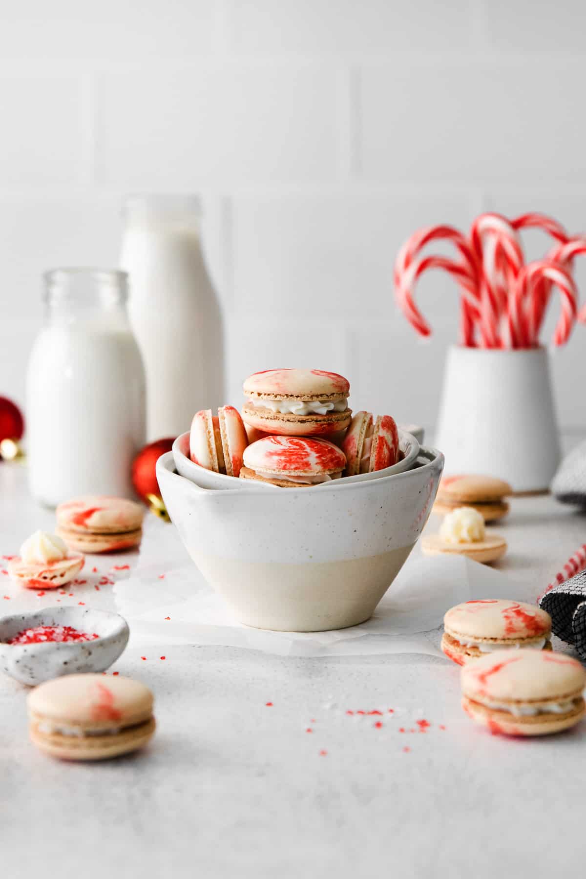 Peppermint macarons in nestled white measuring cups with more of the cookies scattered around and candy canes in the background.