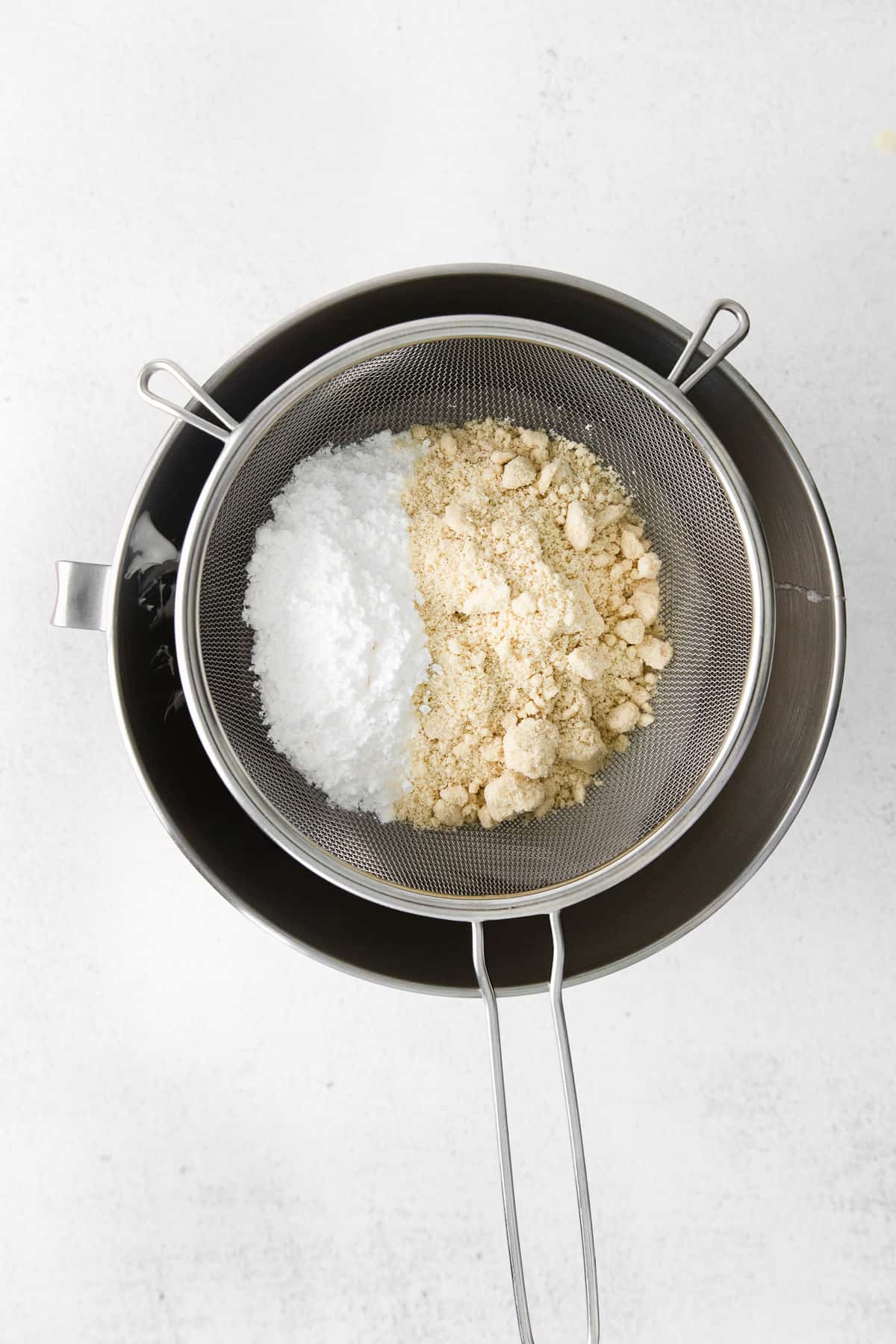 Sifting powdered sugar and almond flour into a mixing bow through a fine mesh sieve.