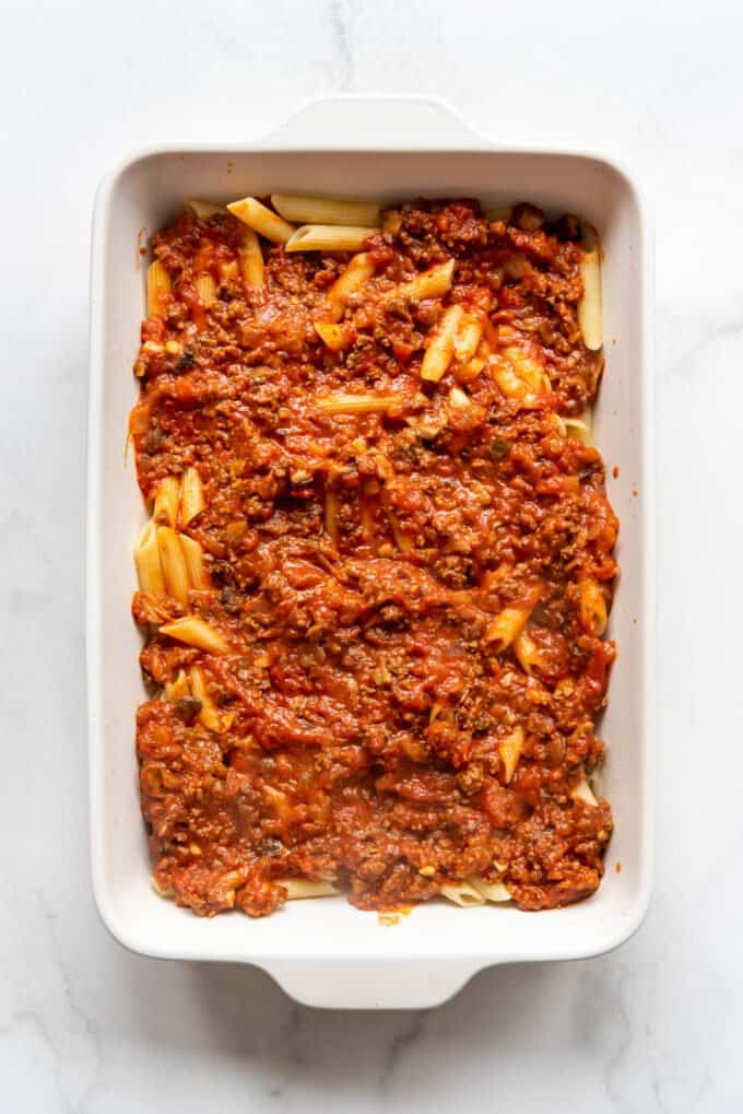 Adding meat sauce on top of cooked penne pasta in a white casserole dish.