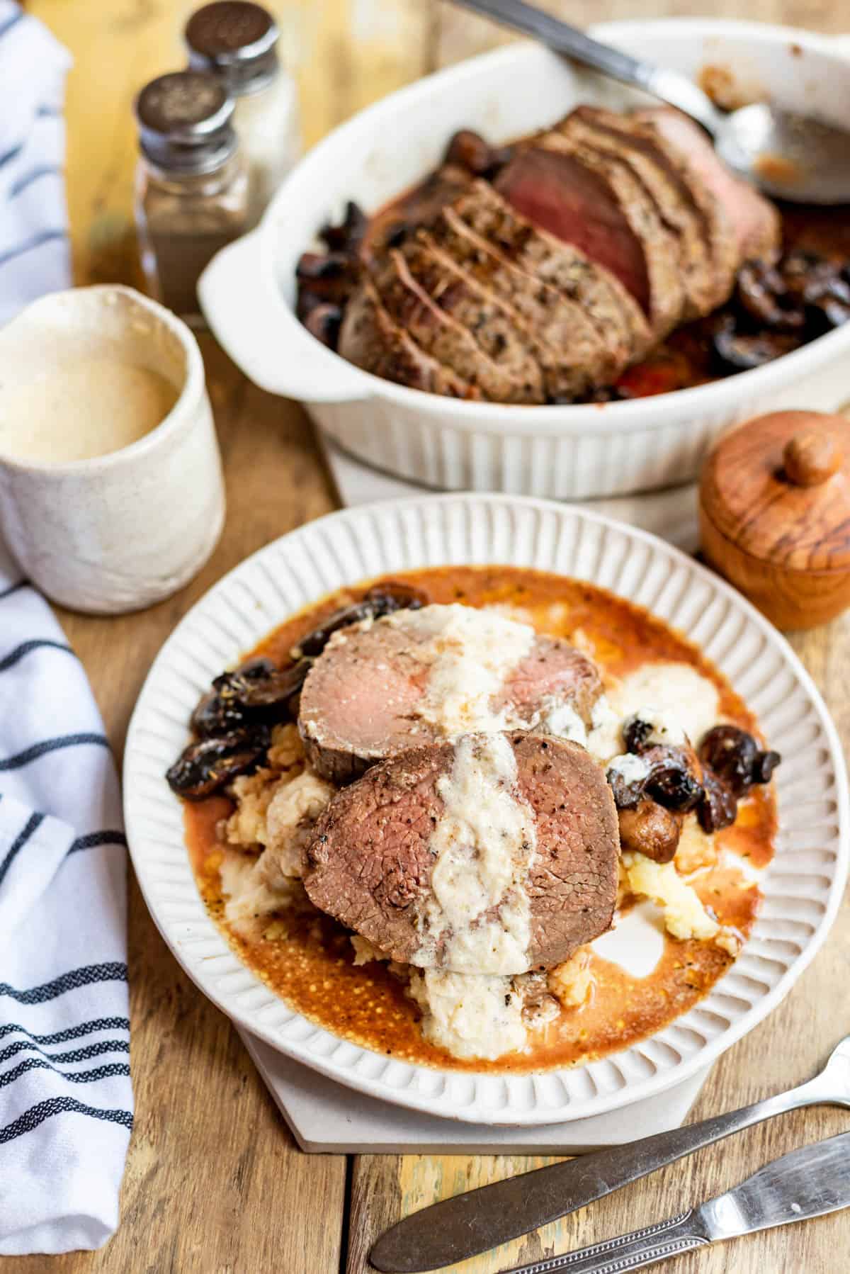 A plate of roasted beef tenderloin with mushrooms and cream sauce in front of the rest of the roast in a white baking dish and cream in a small jar.
