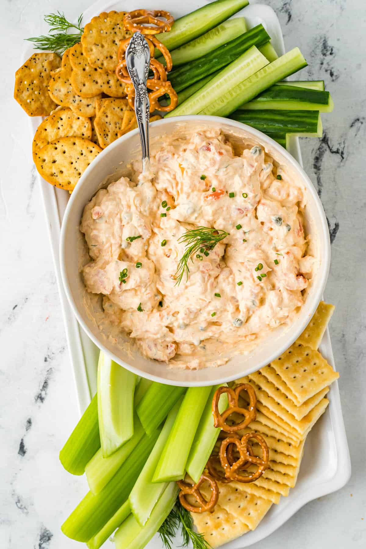 top view of smoked salmon dip in a white bowl with garnish on top and a spoon in it, served in the middle of a platter surrounded by chopped veggies and other snacks. 
