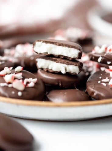 Two halves of a homemade peppermint patty stacked on a plate with other peppermint patties around sprinkled with crushed candy cane pieces.