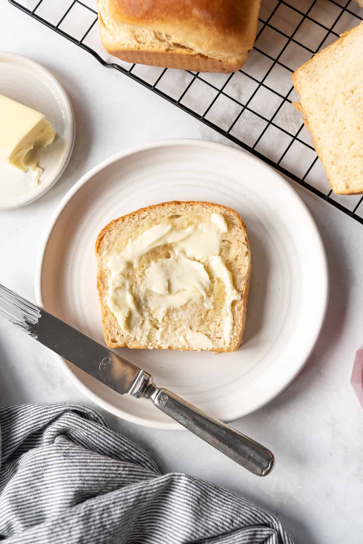 A white plate with a slice of fresh warm homemade bread spread with butter.