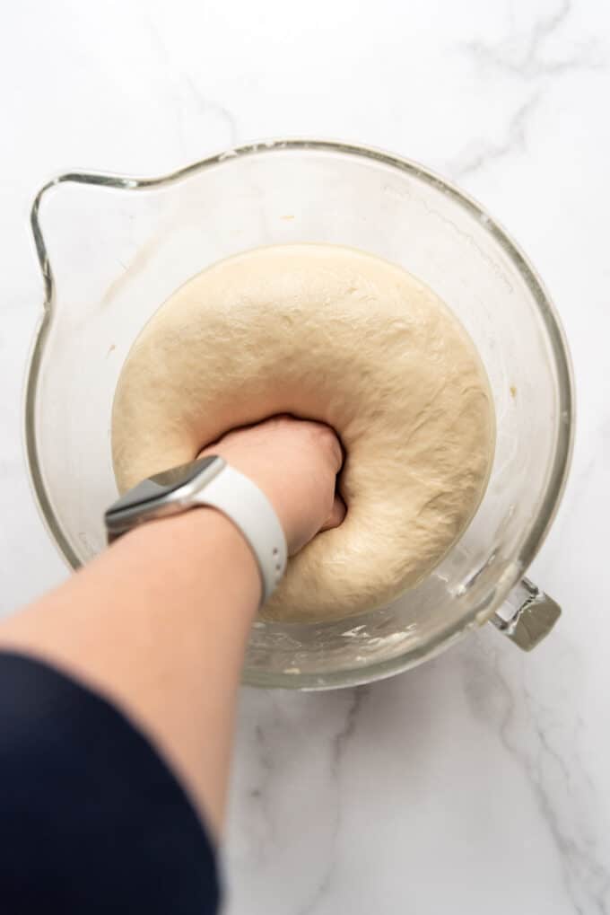 A hand punching down risen bread dough in a glass bowl.