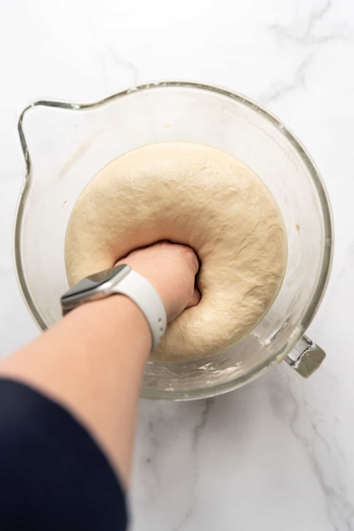 A hand punching down risen bread dough in a glass bowl.