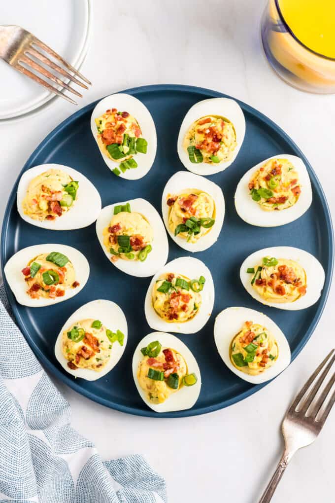 A dozen deviled eggs with bacon and green onions on a blue plate next to forks.