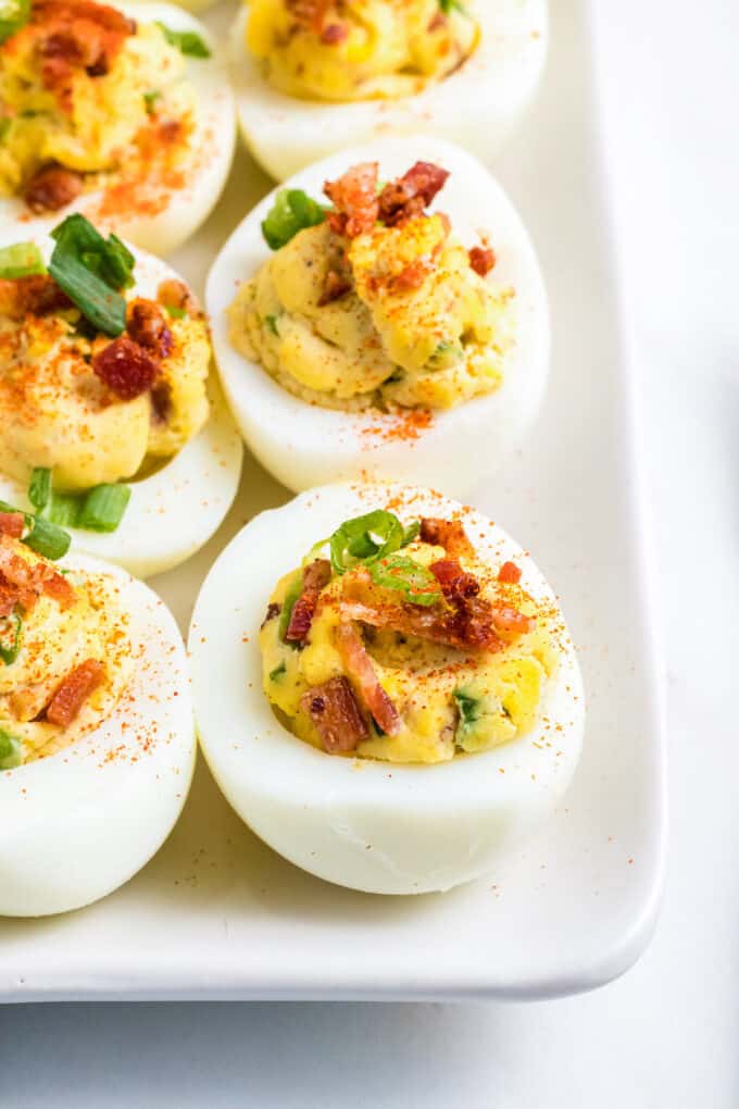 Deviled eggs topped with bacon, green onions, and a sprinkle of paprika on a white plate.