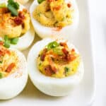 Deviled eggs topped with bacon, green onions, and a sprinkle of paprika on a white plate.