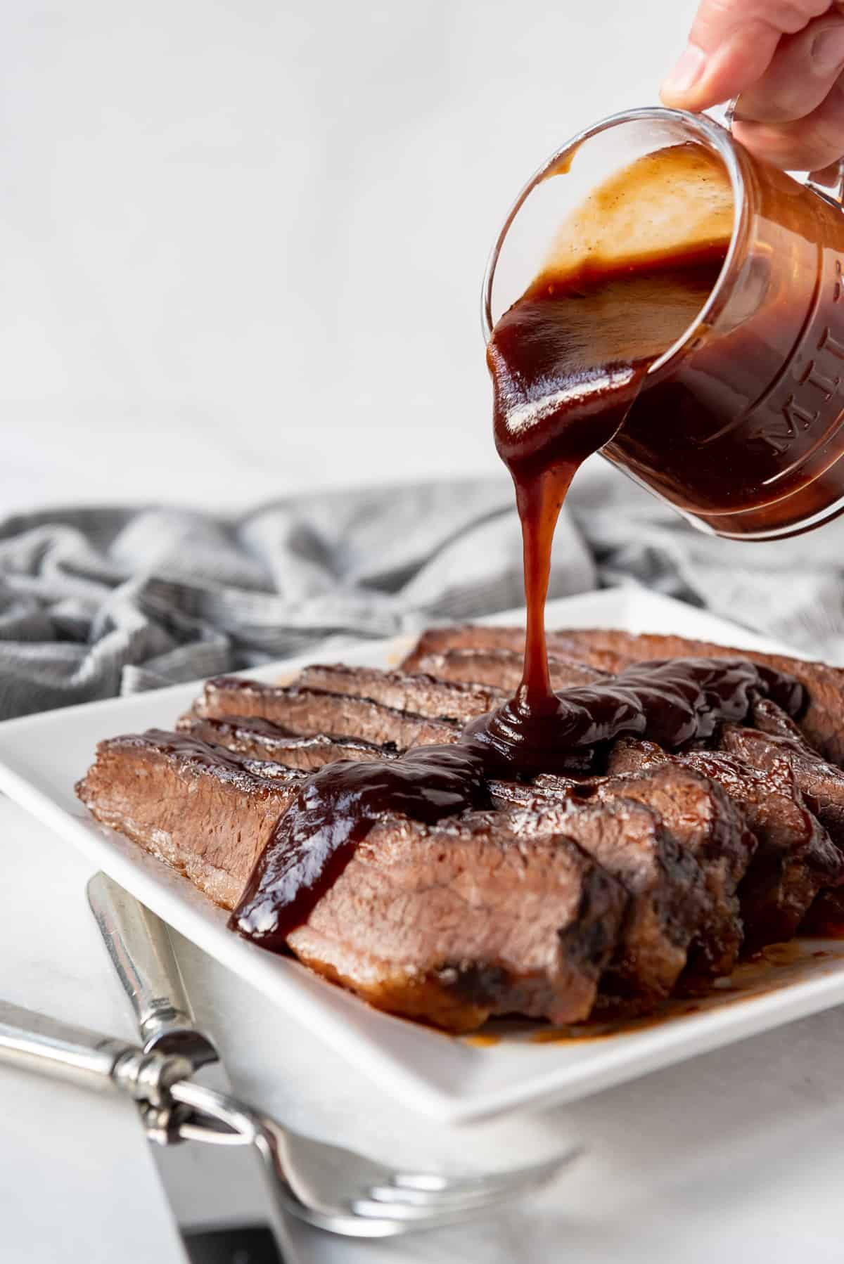 Pouring bbq sauce from a small glass pitcher over sliced beef brisket.