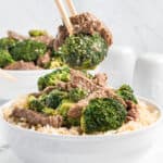 Chopsticks lifting a bite of beef with broccoli from a white bowl.