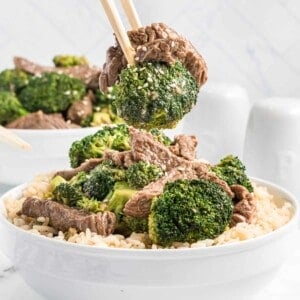 Chopsticks lifting a bite of beef with broccoli from a white bowl.