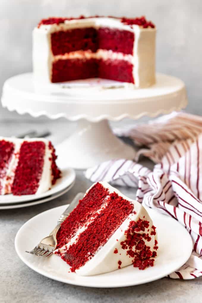 An image of a slice of red velvet cake with cream cheese frosting on a plate with the sliced red velvet layer cake in the background.