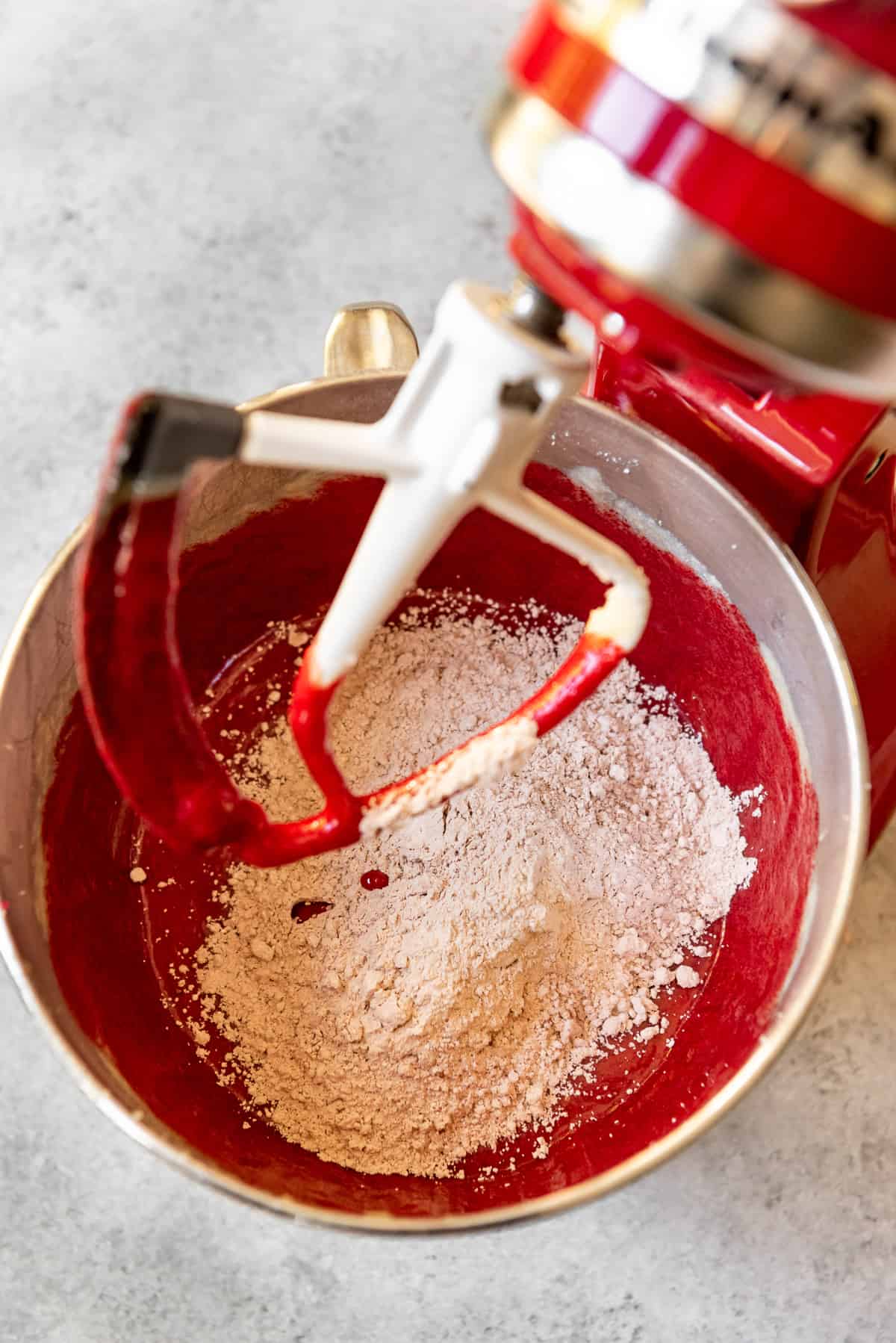 An image of dry ingredients being added to red velvet cake batter.