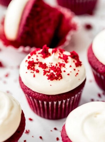 Red velvet cupcakes with frosting on marble counter with red velvet cake crumbs sprinkled on top.