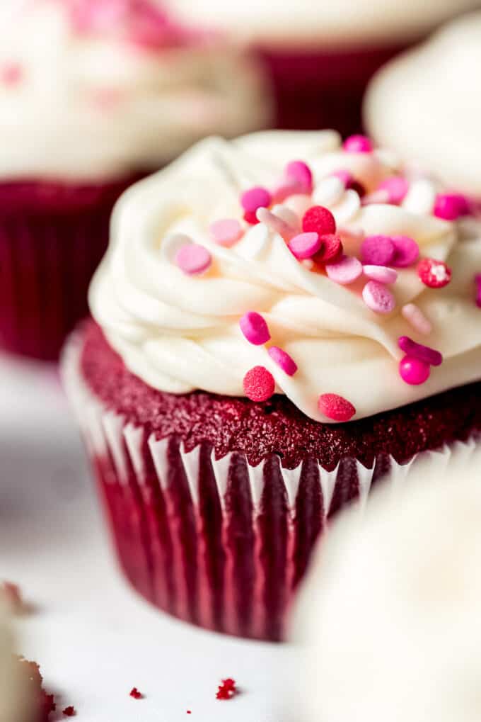 A close image of cream cheese frosting piped onto red velvet cupcakes with pink and red sprinkles.
