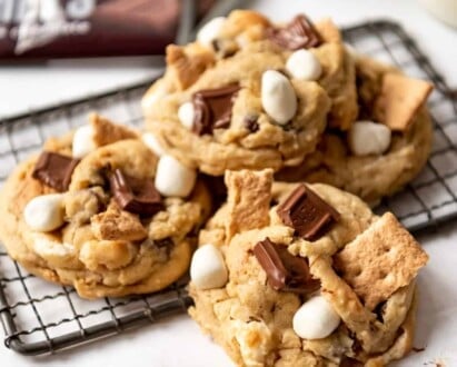 S'mores cookies piled on a wire cooling rack in front of Hershey's bars..